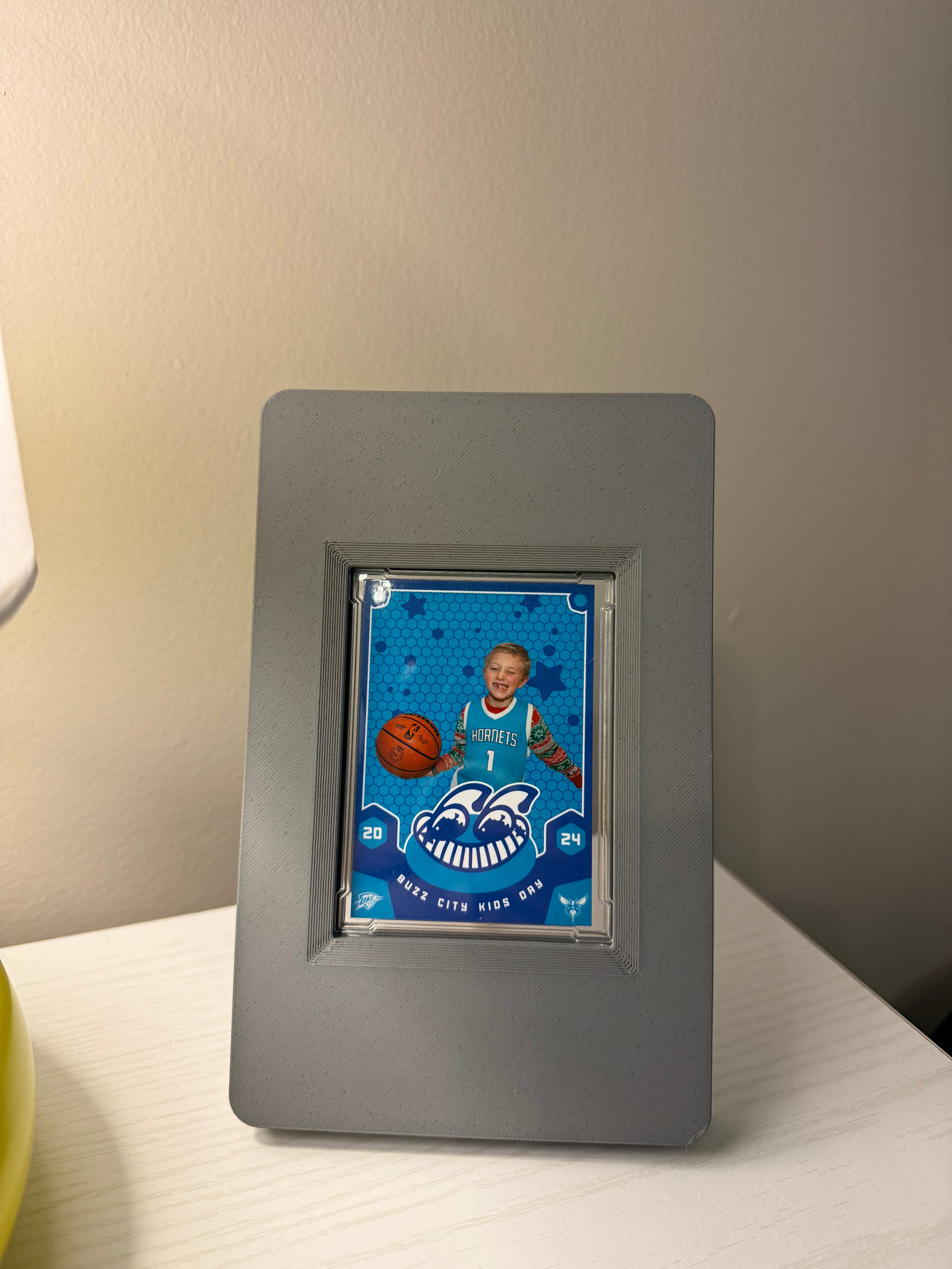 Gray SlabGhost RAW trading card frame with a custom sports card featuring a child with a basketball and team logo on a white surface.