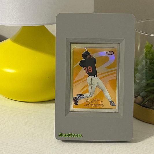Gray SlabGhost graded card frame with a baseball card of Albert Belle on a table next to a yellow vase and plant.